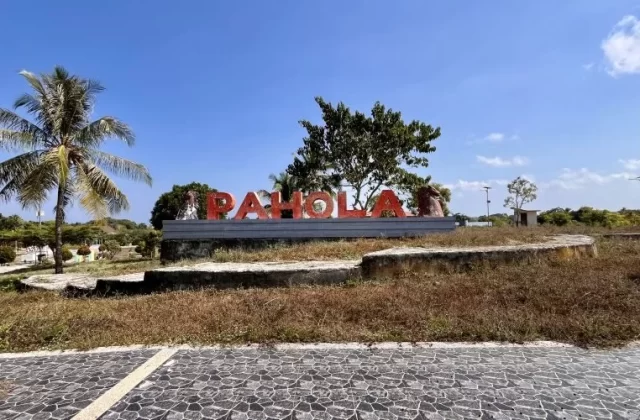 Large red letters spelling "PAHOLA" stand on a concrete base amidst dry grass, palm trees, and other greenery under a clear blue sky.