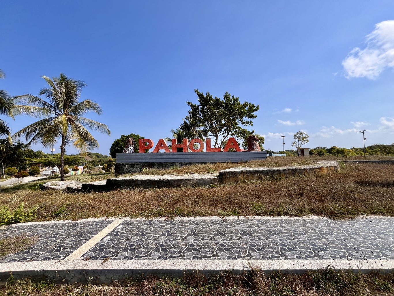 Large red letters spelling "PAHOLA" stand on a concrete base amidst dry grass, palm trees, and other greenery under a clear blue sky.