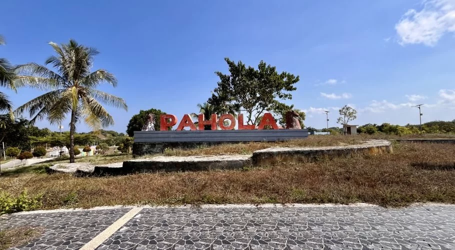 Large red letters spelling "PAHOLA" stand on a concrete base amidst dry grass, palm trees, and other greenery under a clear blue sky.