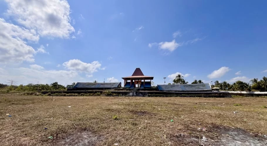 A large, dry grassy field leads to the Pahola Venue, featuring a central building with a traditional red roof flanked by two weathered grandstands under a bright blue, cloudy sky.