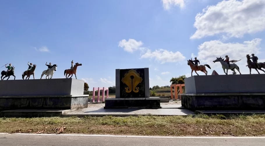 A monument with colorful statues of horsemen on horseback, wielding spears, flanking a central black and gold emblem under a bright blue sky.