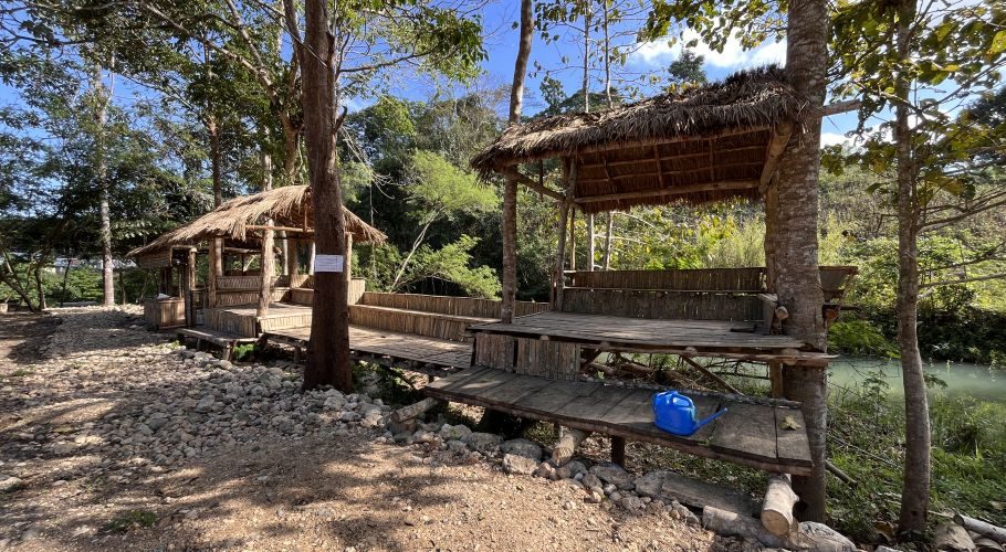 Two rustic, elevated gazebos with thatched roofs and bamboo seating are nestled among lush green trees and rocky ground, beside a light green river under a blue sky.