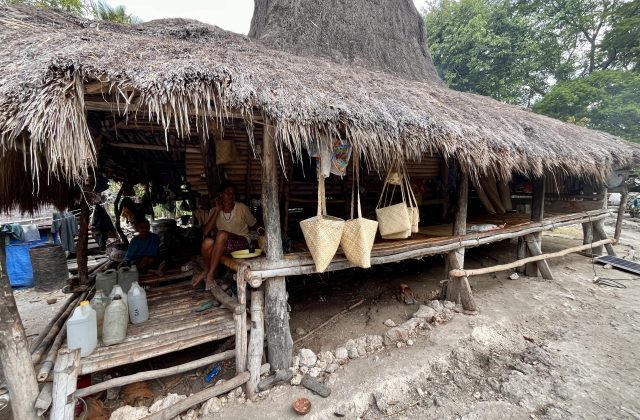 An older woman sits on a raised bamboo platform under a thatched hut, displaying handcrafted pandanus bags for sale, with another person nearby.