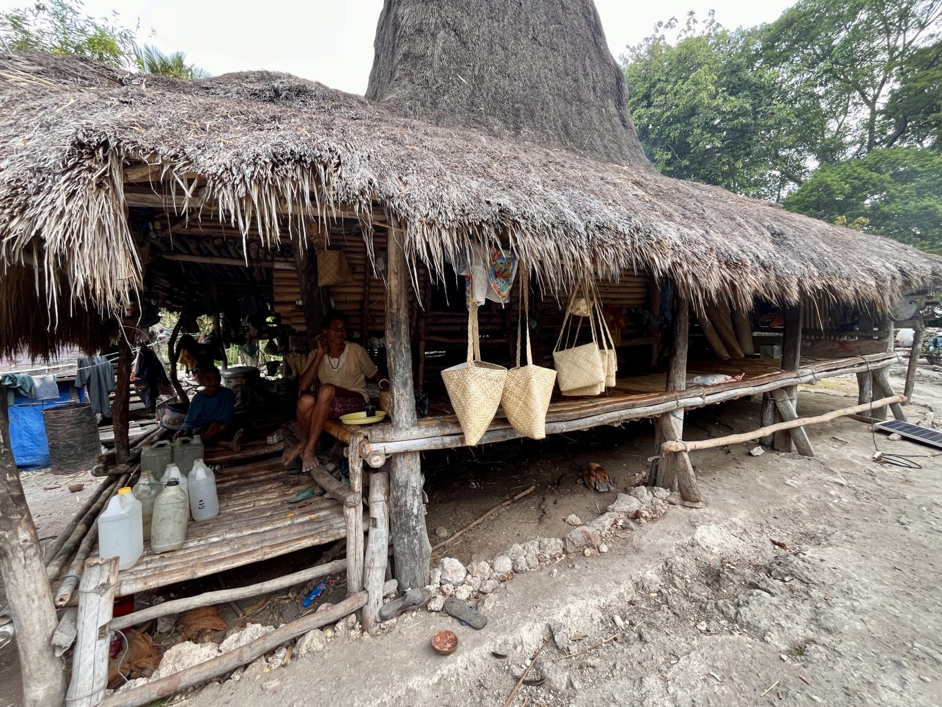 An older woman sits on a raised bamboo platform under a thatched hut, displaying handcrafted pandanus bags for sale, with another person nearby.