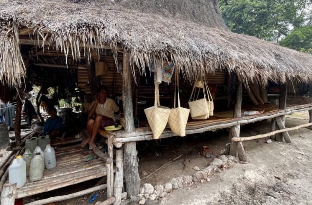 An older woman sits on a raised bamboo platform under a thatched hut, displaying handcrafted pandanus bags for sale, with another person nearby.