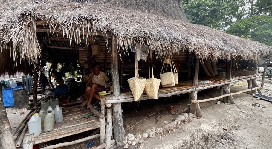 An older woman sits on a raised bamboo platform under a thatched hut, displaying handcrafted pandanus bags for sale, with another person nearby.