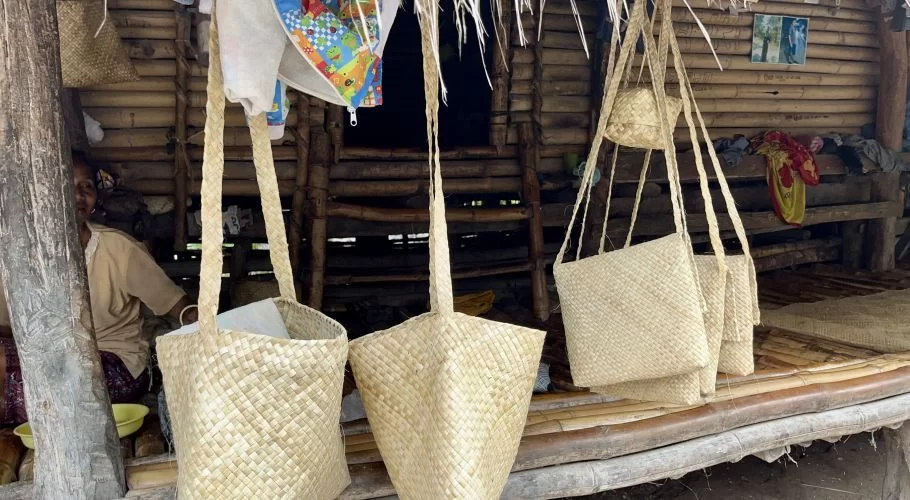 Handcrafted pandanus leaf bags and mats hang from a rustic bamboo stall in Sumba, with a local woman seated inside.