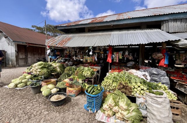 Piles of fresh green vegetables and red tomatoes are displayed at a bustling traditional market stall with a corrugated metal roof under a blue sky.