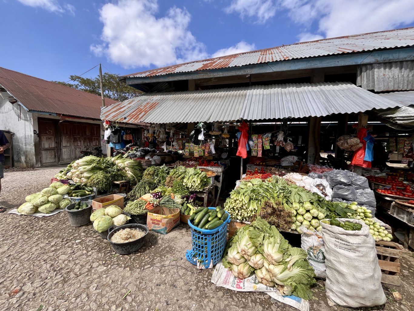 Piles of fresh green vegetables and red tomatoes are displayed at a bustling traditional market stall with a corrugated metal roof under a blue sky.