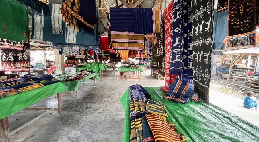 Colorful Sumba ikat textiles hang and are folded on green-covered tables inside a rustic market building. A child sits on the concrete floor.