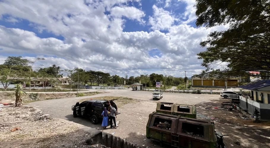 A large, mostly empty dirt parking lot with a black van, two people, and two green dumpsters under a partly cloudy sky. Trees and buildings line the perimeter.