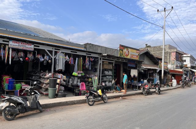 A bustling street scene in Waikabubak, Sumba, shows traditional market stalls displaying household goods and clothing, with several motorbikes parked along the sidewalk under a partly cloudy sky.