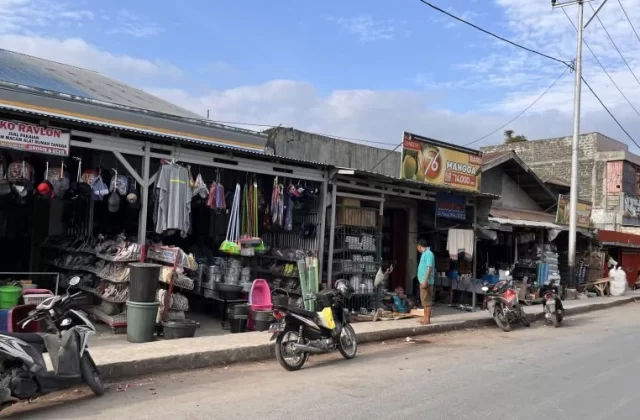 A bustling street scene in Waikabubak, Sumba, shows traditional market stalls displaying household goods and clothing, with several motorbikes parked along the sidewalk under a partly cloudy sky.
