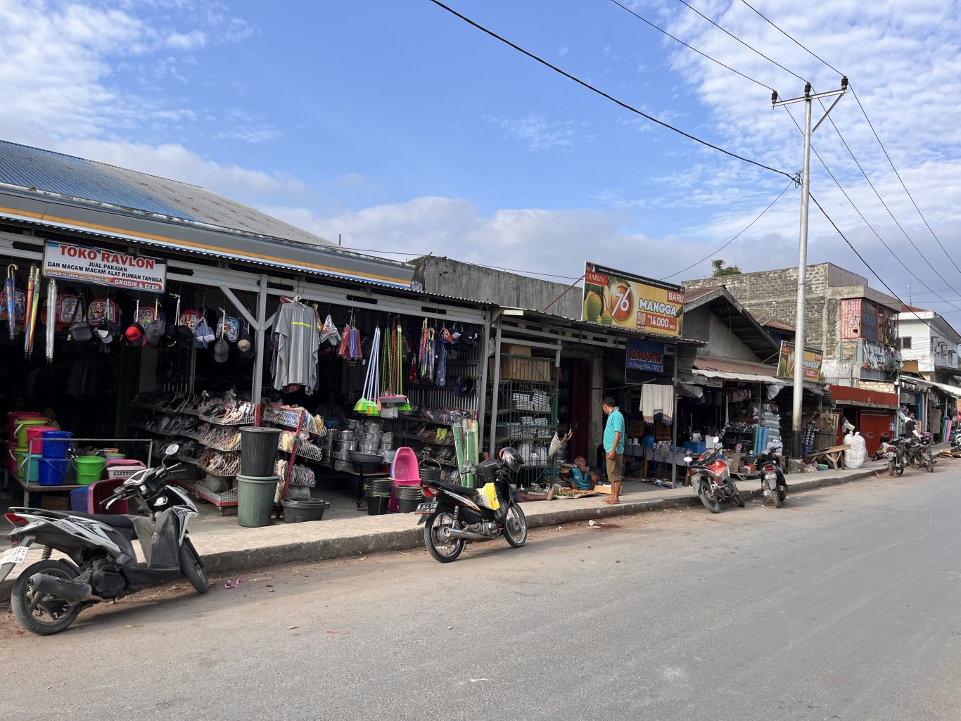 A bustling street scene in Waikabubak, Sumba, shows traditional market stalls displaying household goods and clothing, with several motorbikes parked along the sidewalk under a partly cloudy sky.