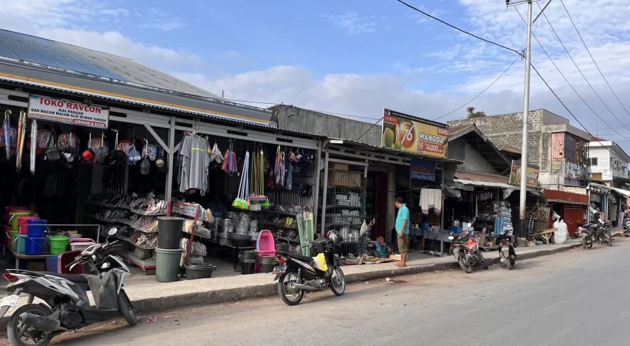 A bustling street scene in Waikabubak, Sumba, shows traditional market stalls displaying household goods and clothing, with several motorbikes parked along the sidewalk under a partly cloudy sky.