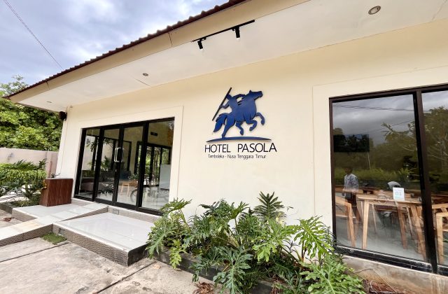 The cream-colored Pasola Hotel exterior features large glass doors, windows, and a blue horse-and-rider logo, with green plants in the foreground.