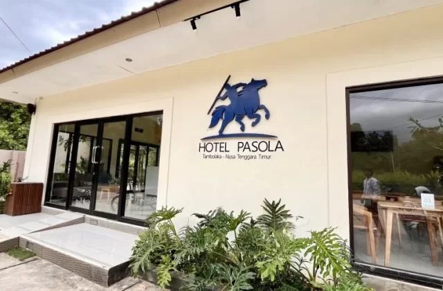 The cream-colored Pasola Hotel exterior features large glass doors, windows, and a blue horse-and-rider logo, with green plants in the foreground.