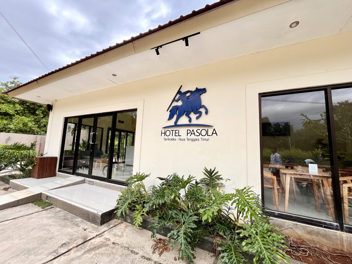 The cream-colored Pasola Hotel exterior features large glass doors, windows, and a blue horse-and-rider logo, with green plants in the foreground.
