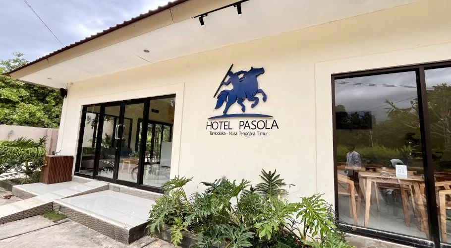 The cream-colored Pasola Hotel exterior features large glass doors, windows, and a blue horse-and-rider logo, with green plants in the foreground.
