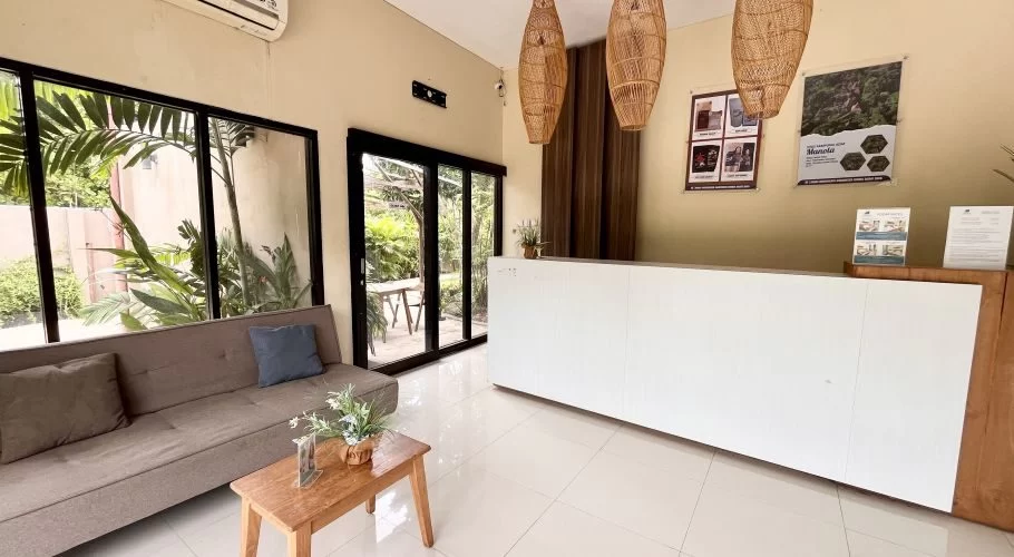A bright hotel lobby features a white reception desk, a beige sofa with pillows, and large windows overlooking a lush green garden.