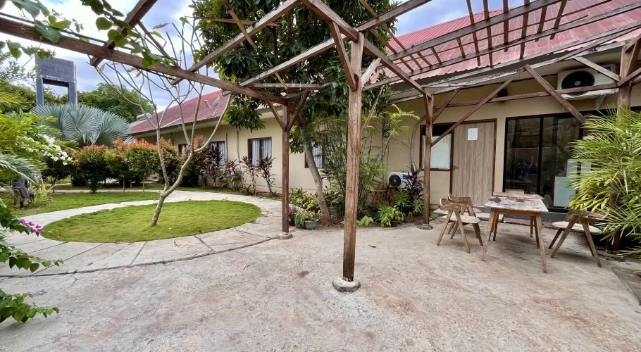 A concrete patio with a wooden pergola and outdoor seating in front of a light-colored, red-roofed hotel building, surrounded by a lush garden under an overcast sky.