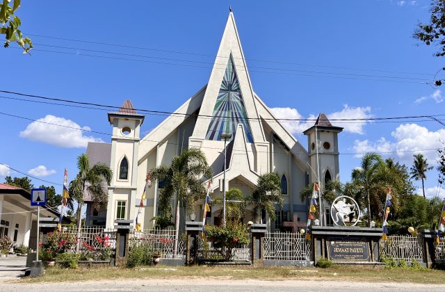 A large, light-colored church with a tall, pointed central spire and two smaller towers stands under a clear blue sky, surrounded by palm trees and a fence with flags.
