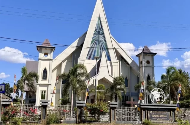 A large, light-colored church with a tall, pointed central spire and two smaller towers stands under a clear blue sky, surrounded by palm trees and a fence with flags.