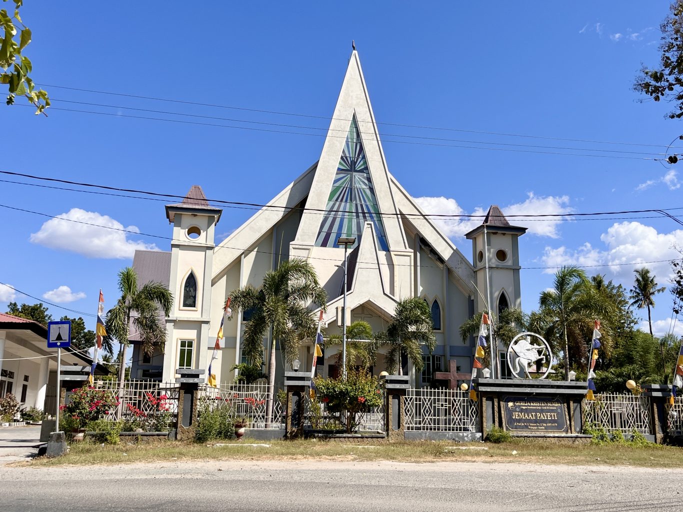 A large, light-colored church with a tall, pointed central spire and two smaller towers stands under a clear blue sky, surrounded by palm trees and a fence with flags.