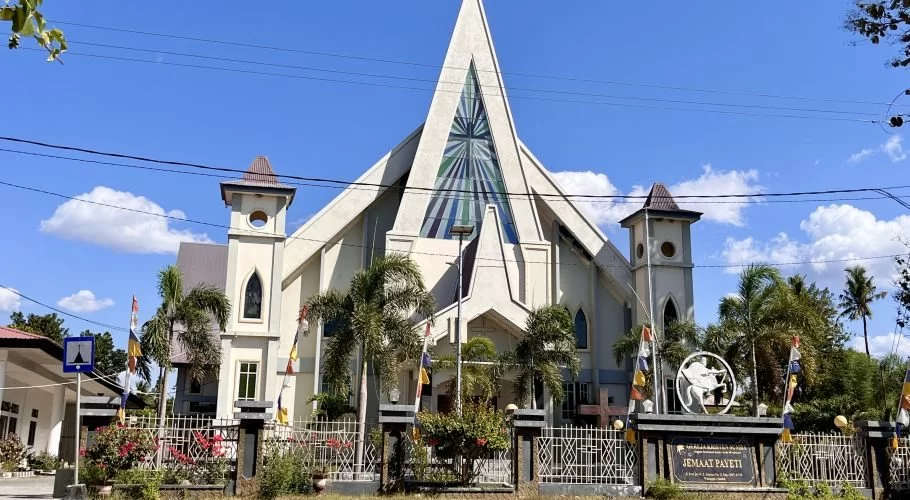 A large, light-colored church with a tall, pointed central spire and two smaller towers stands under a clear blue sky, surrounded by palm trees and a fence with flags.