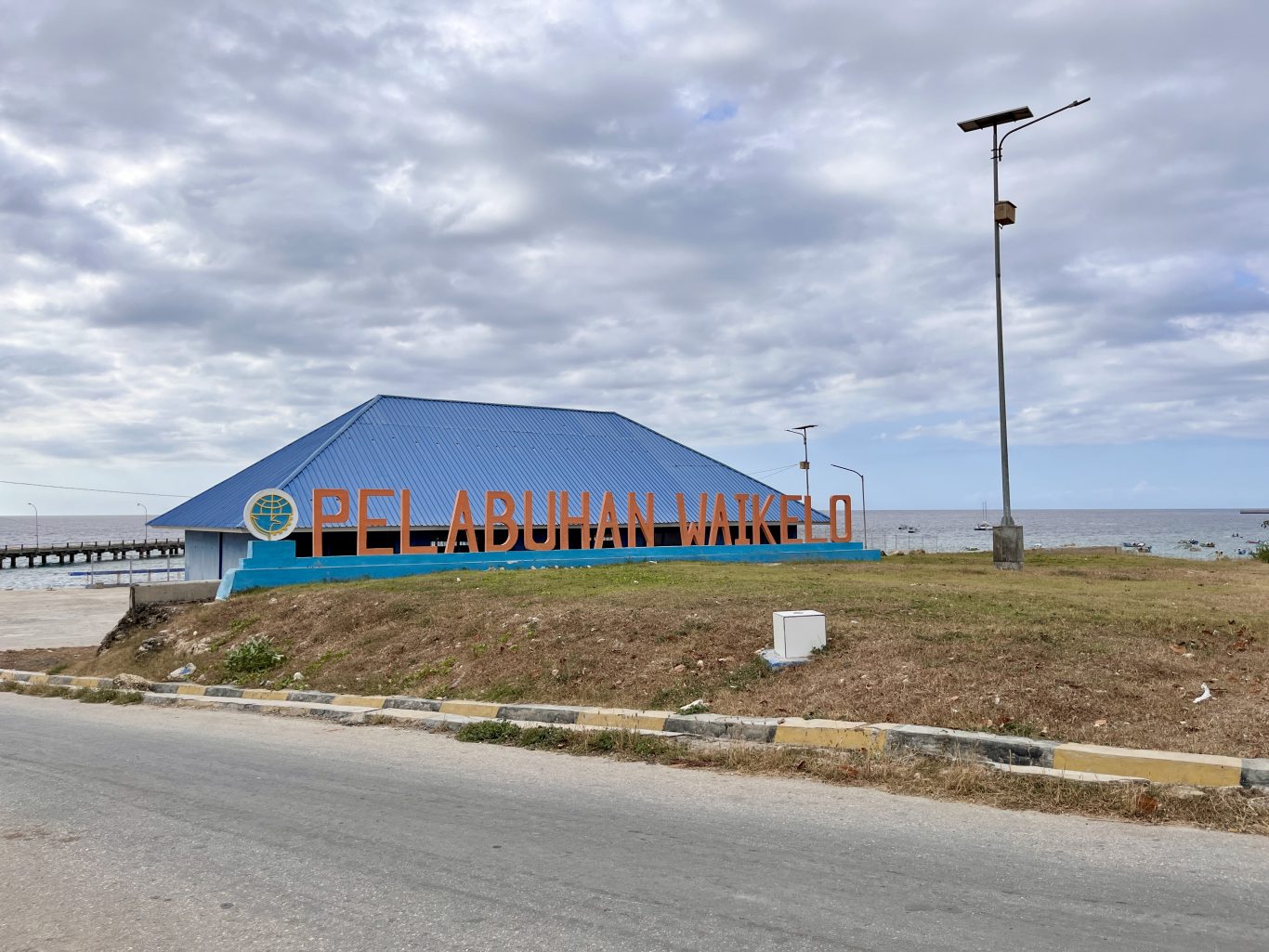 The Pelabuhan Waikelo port entrance features a blue-roofed building and large orange letters, with a pier and small boats on the sea under a cloudy sky.
