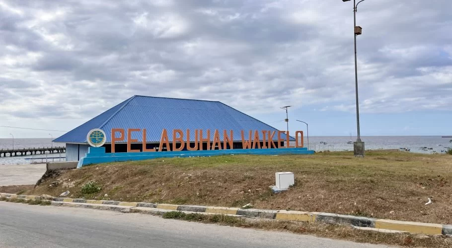 The Pelabuhan Waikelo port entrance features a blue-roofed building and large orange letters, with a pier and small boats on the sea under a cloudy sky.
