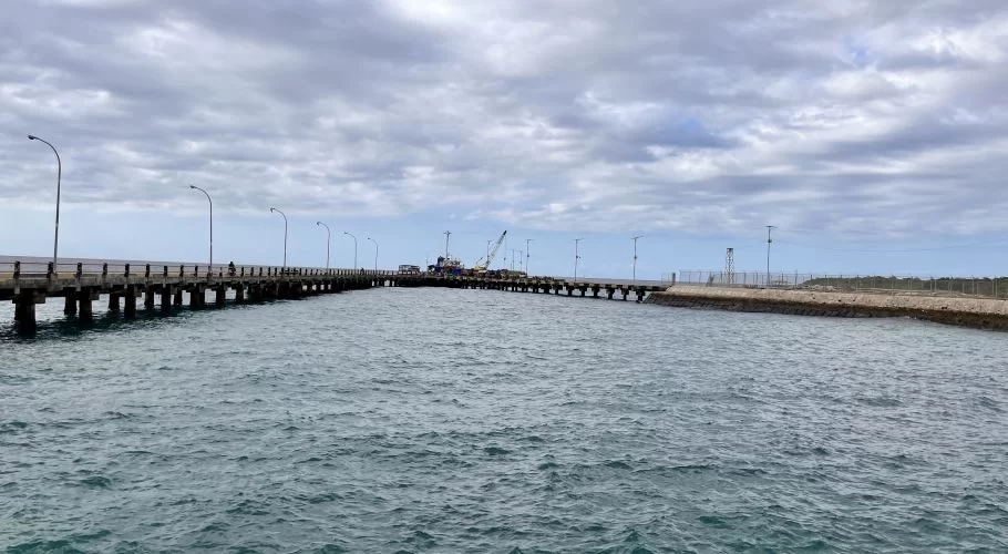 A long concrete pier with streetlights extends into choppy blue-green water under a cloudy sky. Cranes and vehicles are visible at the pier's end.