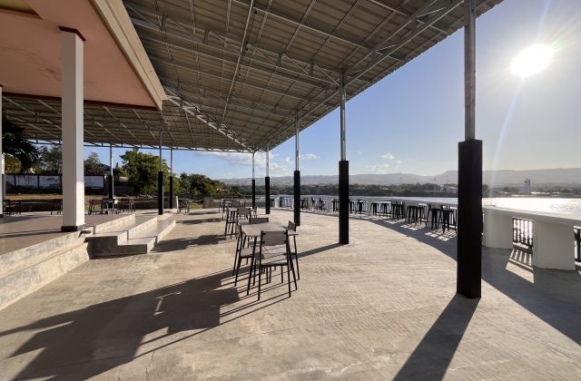 A large, covered outdoor dining terrace at Permata Restaurant overlooks the sunny port and ocean in Waingapu, Sumba, with tables and chairs.