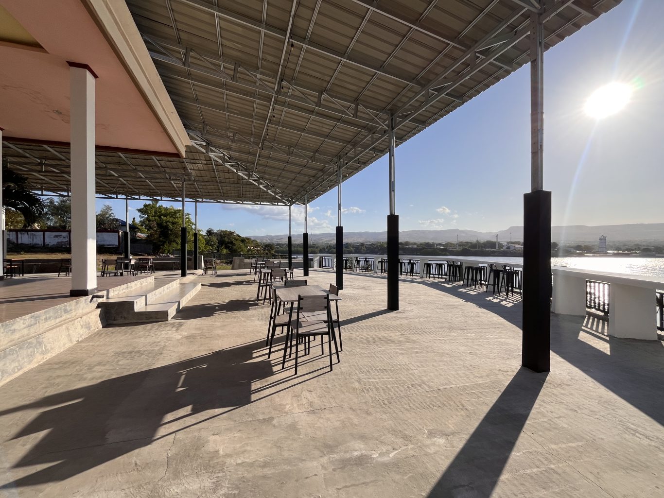 A large, covered outdoor dining terrace at Permata Restaurant overlooks the sunny port and ocean in Waingapu, Sumba, with tables and chairs.