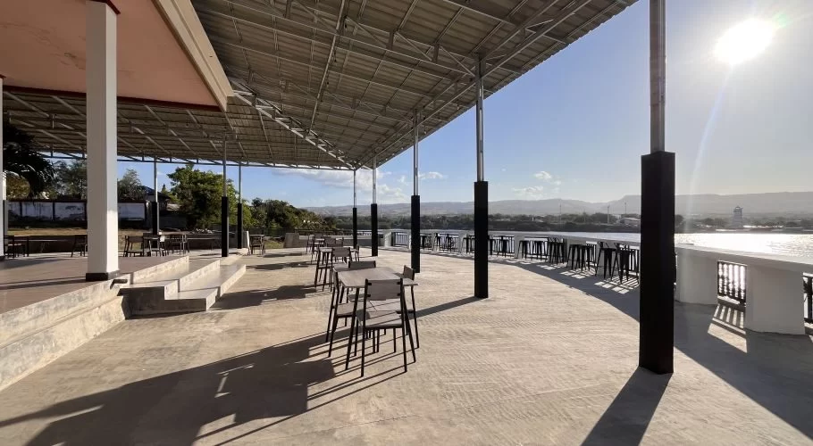 A large, covered outdoor dining terrace at Permata Restaurant overlooks the sunny port and ocean in Waingapu, Sumba, with tables and chairs.