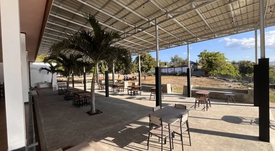 Outdoor restaurant patio with numerous empty tables and chairs under a metal roof, featuring palm trees and a view of a dry open field under a blue sky.