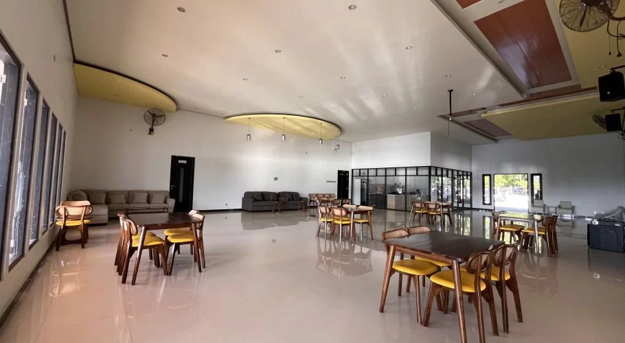 A spacious, modern restaurant interior features light tiled floors, white walls, dark wooden tables with yellow chairs, and comfortable sofas.
