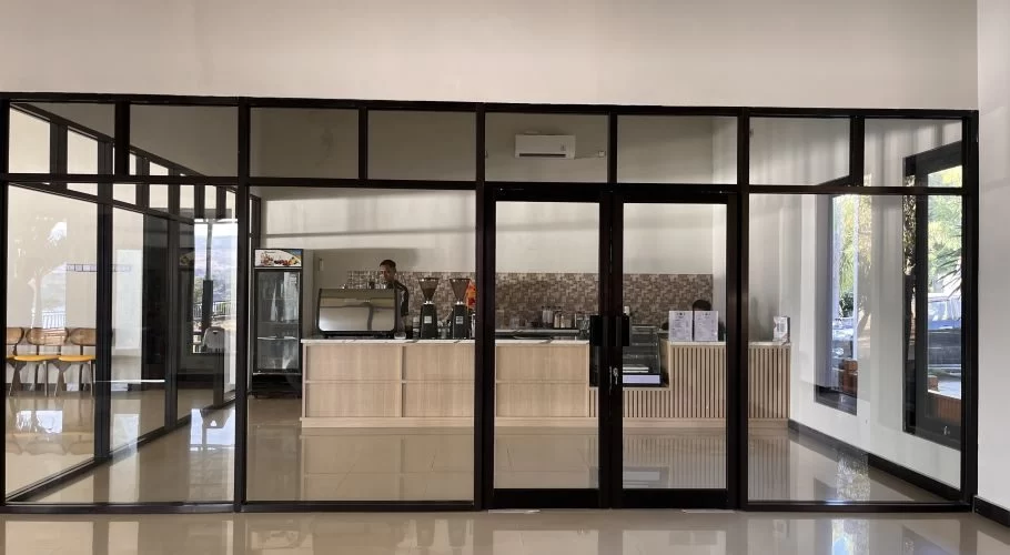 A modern cafe interior features a barista behind a light wood counter with coffee machines, framed by black glass partitions and doors.