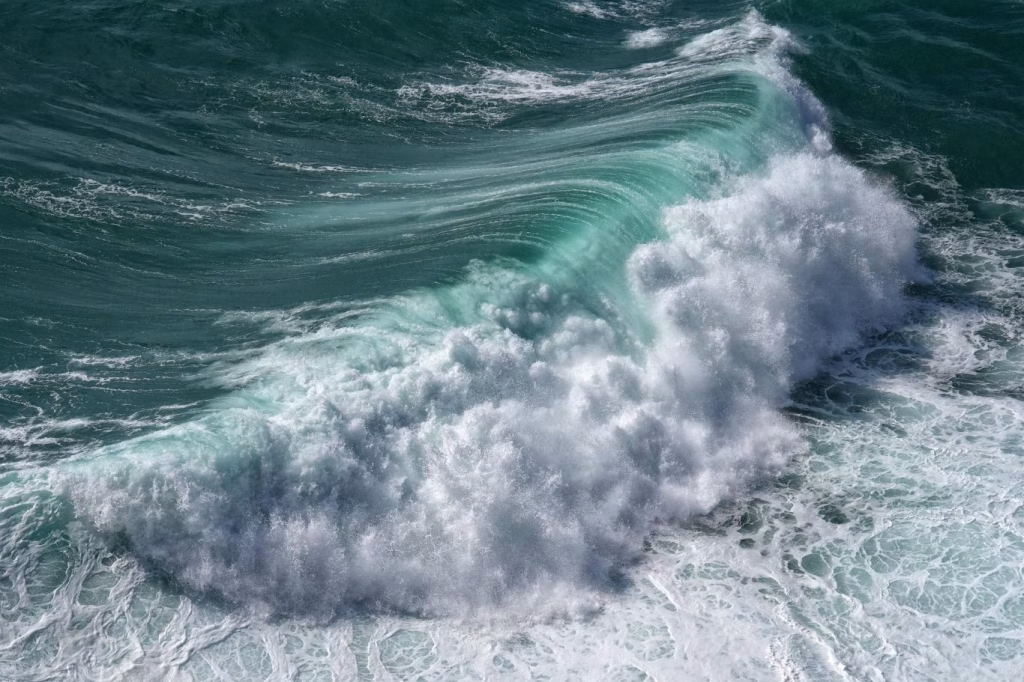 A powerful, teal-colored ocean wave curls and breaks, creating a large mass of white foam and spray across the water's surface.
