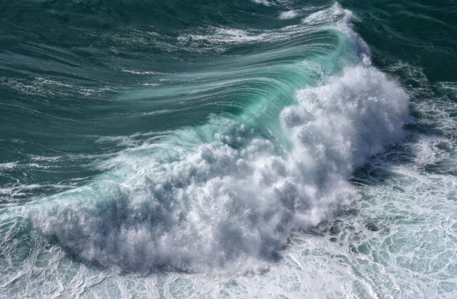 A powerful, teal-colored ocean wave curls and breaks, creating a large mass of white foam and spray across the water's surface.