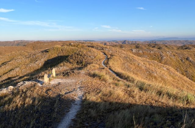 Rolling golden-brown hills covered in dry grass stretch under a clear blue sky, with a winding dirt path and two stone structures in the foreground.