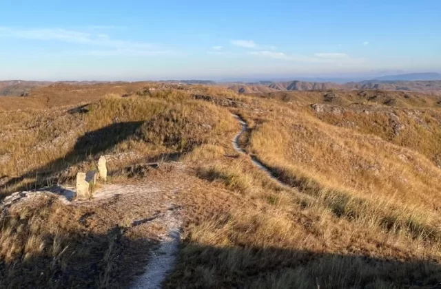 Rolling golden-brown hills covered in dry grass stretch under a clear blue sky, with a winding dirt path and two stone structures in the foreground.