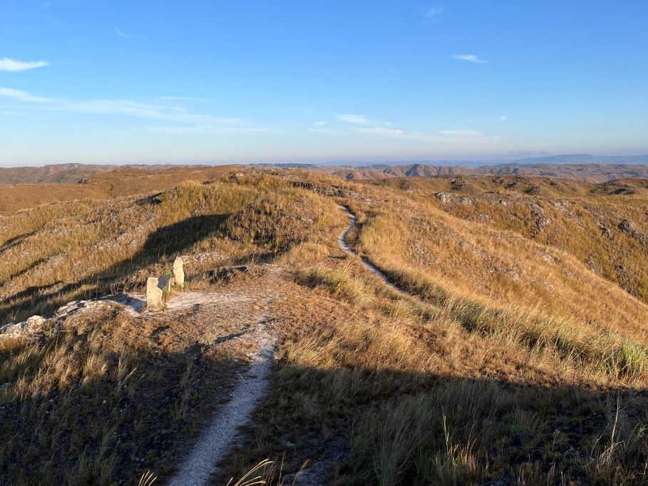 Rolling golden-brown hills covered in dry grass stretch under a clear blue sky, with a winding dirt path and two stone structures in the foreground.