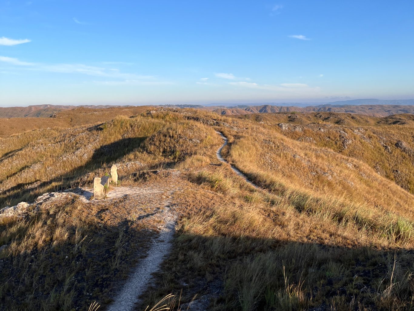 Rolling golden-brown hills covered in dry grass stretch under a clear blue sky, with a winding dirt path and two stone structures in the foreground.