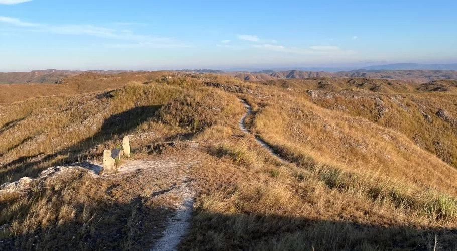 Rolling golden-brown hills covered in dry grass stretch under a clear blue sky, with a winding dirt path and two stone structures in the foreground.