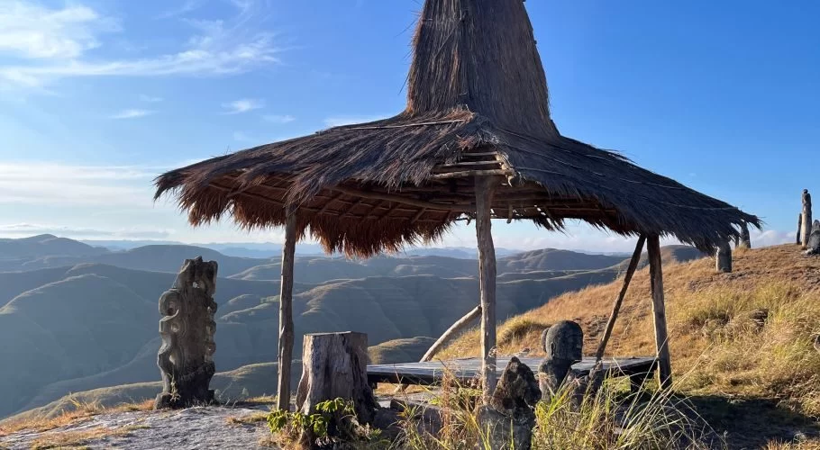 A thatched shelter and megalithic statues on a golden grassy hilltop overlook rolling blue-grey hills under a clear sky.