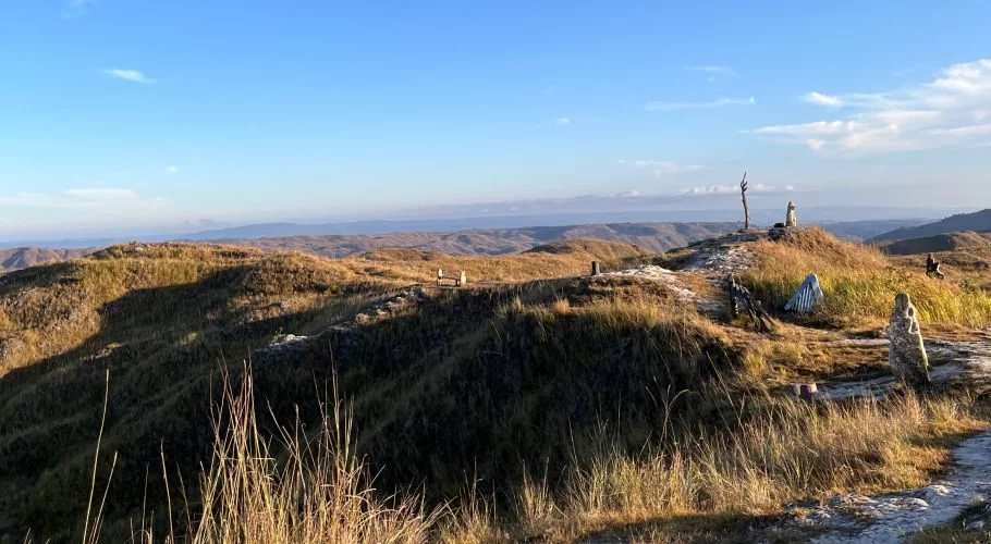 Golden-brown grassy hills roll under a clear blue sky, with ancient megalithic stones and a wooden structure visible on the sunlit landscape.