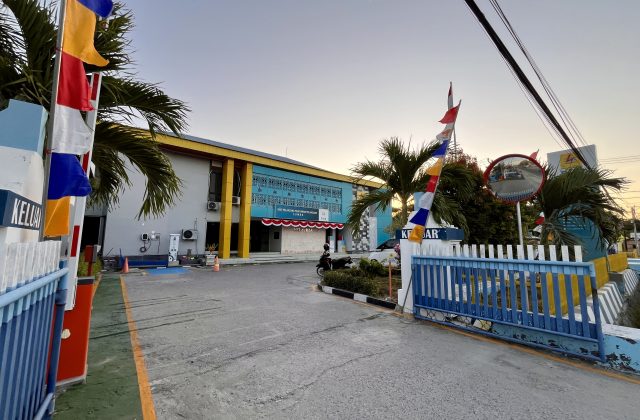 The PLN Office in Waingapu features a blue and yellow building with a driveway, blue and white gates, colorful flags, and palm trees under a clear sky.