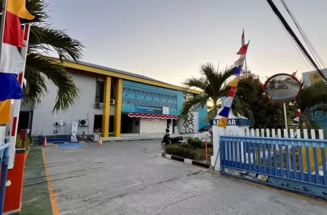 The PLN Office in Waingapu features a blue and yellow building with a driveway, blue and white gates, colorful flags, and palm trees under a clear sky.