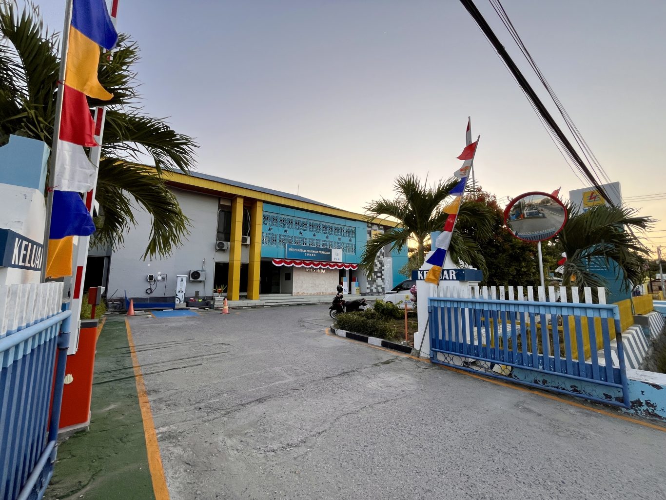 The PLN Office in Waingapu features a blue and yellow building with a driveway, blue and white gates, colorful flags, and palm trees under a clear sky.